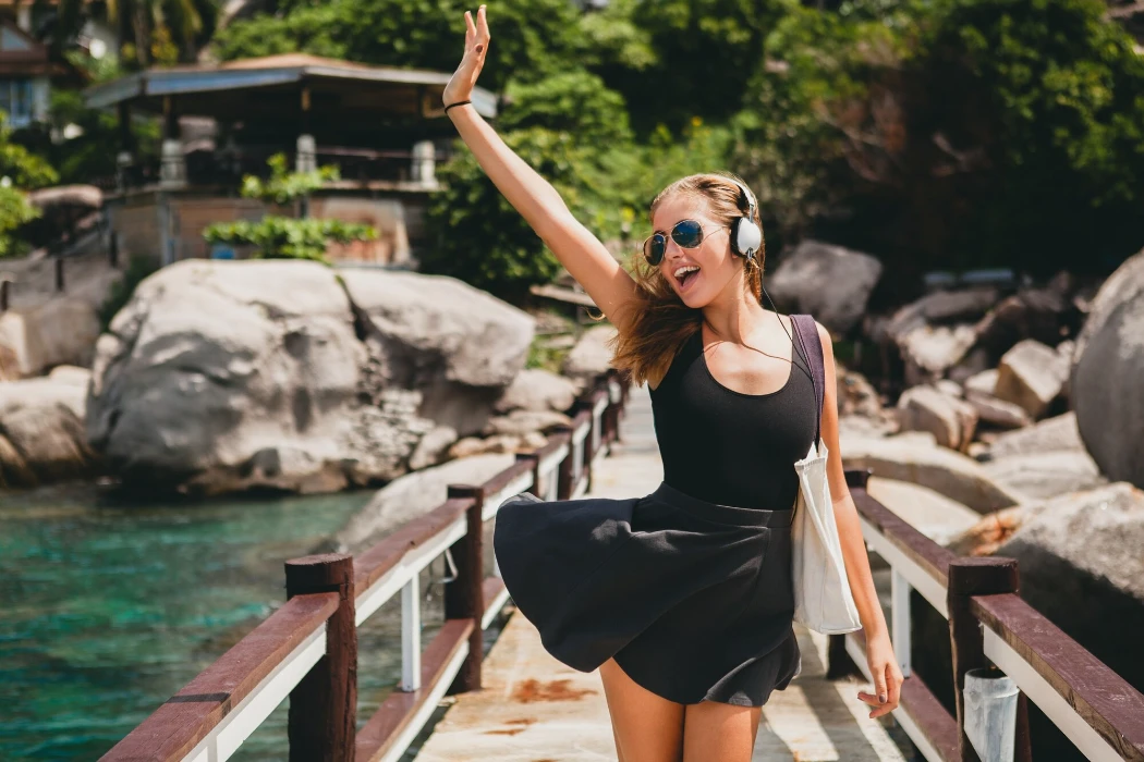 Young stylish hipster woman positive mood, smiling happy, traveling around world, sunglasses, headphones, listening to music, tropical summer vacation, skirt, enjoy freedom, flirty, sexy